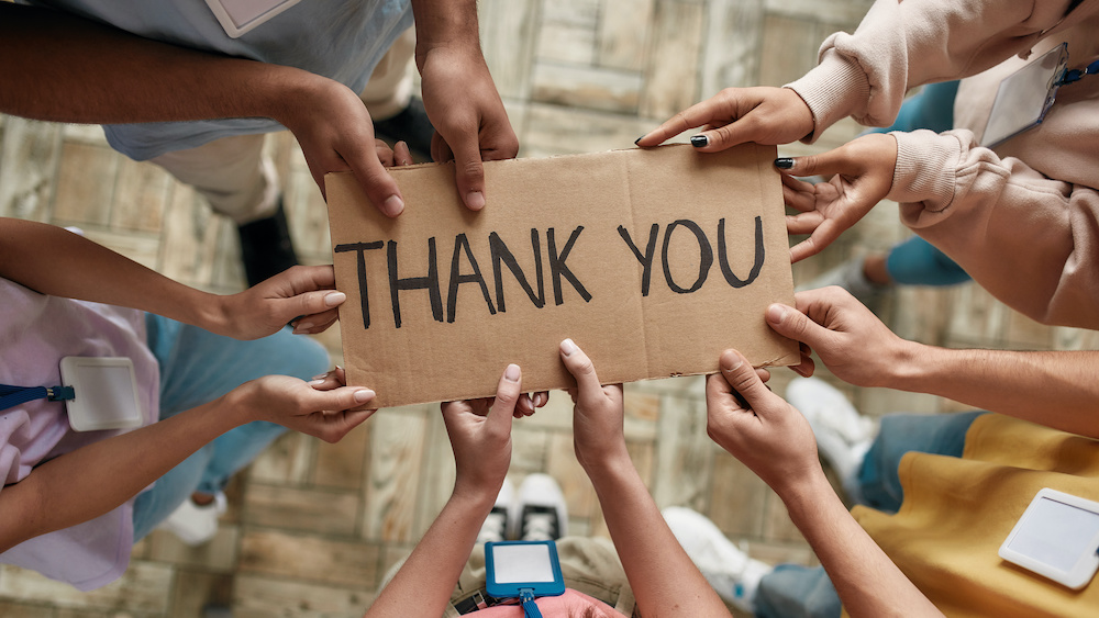 Top view of diverse young volunteers holding card with Thank you lettering while standing in charitable organization office, Selective focus on hands and inscription, Web Banner